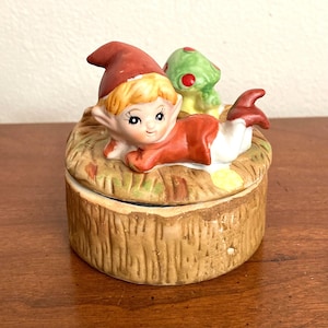 May include: A ceramic trinket box featuring a whimsical elf figure in a red outfit and hat, lying on a bed of straw. A green mushroom with red spots sits nearby. The box has a textured, wood-like appearance.