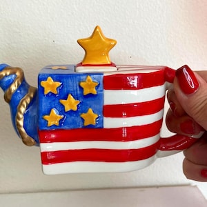 May include: A patriotic teapot featuring the American flag design. The teapot has a blue spout with gold accents, a blue section with yellow stars, and red and white stripes. The lid is topped with a yellow star.