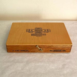 May include: A rectangular light brown wooden cigar box with a brass latch. The lid features black text reading IMPORTED PUROS INDIOS CIGARS MADE IN HONDURAS. The front edge displays the text 25 TORO ESPECIAL and PUROS INDIOS CIGARS.