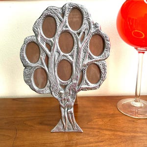 May include: A silver-colored tree-shaped picture frame with eight oval openings. The frame features a detailed design of a tree with figures at the base. A red glass is visible in the background. The frame is made of metal and sits on a wooden surface.