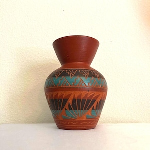 May include: A terracotta-colored vase with a flared top and a rounded body. The vase features a geometric design in turquoise, black, and orange. The design includes triangles and fan-like shapes, adding a decorative touch to the ceramic piece.
