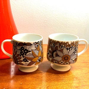 May include: Two white ceramic mugs with a brown, gold, and black floral pattern. The mugs have a pedestal base.