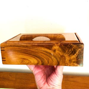 May include: A wooden box with a lid and a handle. The box is a warm brown color with a visible wood grain pattern. The handle is a curved cutout in the center of the lid. The box is held in a person's hand.