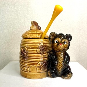 May include: A ceramic honey pot shaped like a beehive with a lid, featuring embossed bees and a small bear figurine. The honey dipper is yellow. The pot is brown and tan.