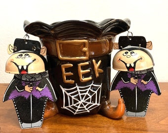 May include: A brown ceramic witch's hat with a spiderweb design and the word "EEK" on the front. Two black and purple painted ceramic vampire bats with fangs hang from the hat.