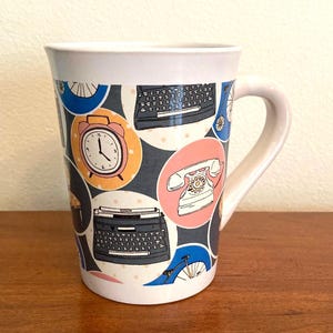 May include: White ceramic mug with a wrap-around design featuring retro illustrations of a typewriter, alarm clock, rotary phone, and bicycle wheel. The design is set against a dark gray background with white polka dots and colorful circular frames.