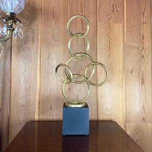 Vintage abstract 21.5” metal sculpture, geometric interlocking rings, textured gold metallic finish, wood base, Minimalist tabletop decor.
