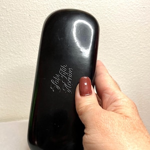 May include: A black, rectangular eyeglasses case with rounded edges. The case has the words "Saks Fifth Avenue" in a cursive font. The case is being held by a person.