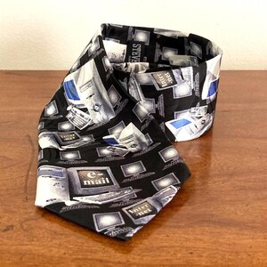 May include: Black necktie with a repeating pattern of computer monitors, keyboards, and the words "e-mail" and "Internet" in white. The tie is made of a shiny fabric and is folded on a wooden surface.
