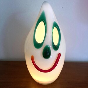 May include: A whimsical, teardrop-shaped lamp with a smiling face design. The lamp features a white body with green eye and nose accents, and a red and orange smiling mouth. The eyes and the body of the lamp glow with a warm light.