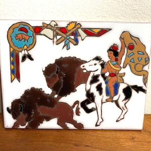 May include: A white ceramic tile with a colorful Native American scene. A Native American person rides a white horse with black spots, holding a brown and yellow blanket. Two brown bison are in the background. The tile has a red, yellow, and blue border with a Native American symbol in the corner.