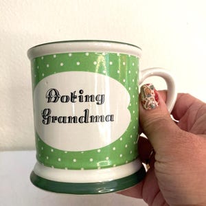 May include: A ceramic mug with a green polka dot pattern and the words "Doting Grandma" in black script. The mug has a white oval shape in the center where the text is displayed. The handle is white and the rim and base are green.