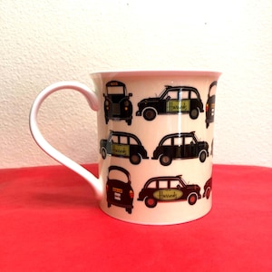 May include: A white ceramic mug featuring a repeating pattern of black London taxis. The taxis have the word "Harrods" on the side. The mug has a white handle and is set against a red surface.