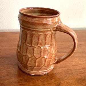 May include: A handmade ceramic mug with a textured, honeycomb-like pattern in shades of brown. The mug has a sturdy handle and a slightly flared rim. The mug is suitable for hot or cold beverages.