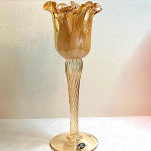 May include: A tall, amber-colored glass vase with a tulip-shaped top and a twisted stem. The vase has a wide, circular base and a label near the bottom. The glass has a glossy finish, reflecting light. The overall design is elegant and decorative.