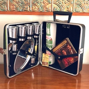 May include: Open vintage black cocktail case with silver metal cups, a serving tray, and a bottle of Squirt. The case also contains a book titled "History of Mixed Drinks" and a small tag.