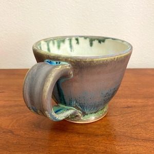 May include: A handmade ceramic mug with a unique glaze featuring shades of brown, blue, and green. The mug has a wide, rounded shape and a curved handle.