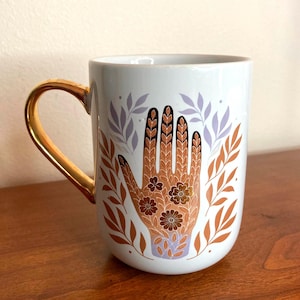May include: White ceramic mug with a gold handle. The mug features a detailed illustration of a hand with floral and leaf designs in shades of brown, purple, and gold. The design is set against a backdrop of purple and orange leaves.