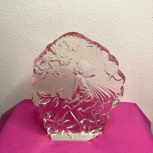 May include: A clear glass sculpture featuring a fairy figure amidst floral and leaf details. The fairy has wings and is looking upwards. The sculpture has a textured, irregular edge and sits on a pink surface.