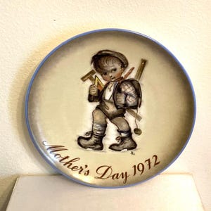 May include: A decorative plate with a blue rim and a central illustration of a young boy carrying tools and a backpack. The plate is inscribed with "Mother's Day 1972" in brown script. The boy is wearing a hat, overalls, and boots.