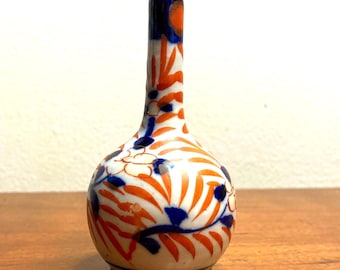 May include: A small, white ceramic vase with a bulbous body and a narrow neck. The vase is decorated with a floral pattern in shades of orange, blue, and gold.