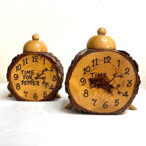 May include: Two wooden salt and pepper shakers. Each shaker is shaped like a clock face with the words "TIME FOR PEPPER" and "TIME FOR SALT" on the face. The shakers are made of light brown wood with a bark edge.