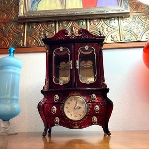 May include: A red wooden jewelry box with a clock face and three drawers. The box has a glass front with a gold trim and the words "Lord is Home in the Morn" written on the inside.