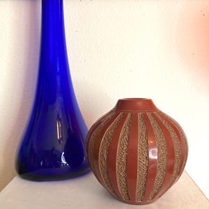 May include: A tall, cobalt blue glass vase stands next to a small, round, reddish-brown vase. The round vase has vertical stripes of a lighter brown, textured material. Both vases are on a white surface.