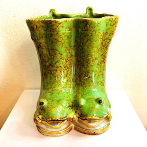 May include: A ceramic planter shaped like a pair of rain boots in a bright green glaze with mottled brown accents. The front of the boots features molded frog faces with black eyes and wide mouths. The top opening has a wavy, uneven rim design.