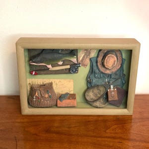 May include: A framed relief sculpture depicting fishing gear. The artwork includes a fish, fishing rod, tackle box, vest, hat, and creel. The color palette features greens, browns, and tans, with a wooden frame.