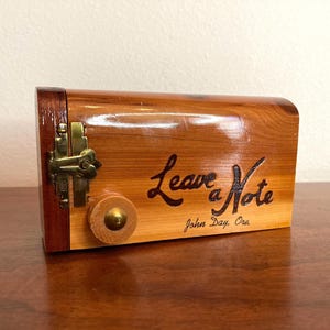 May include: A wooden box with a hinged lid and a brass clasp. The box is made of light and dark brown wood and has the words "Leave a Note" engraved on the front. The text "John Day, Ore" is also engraved.