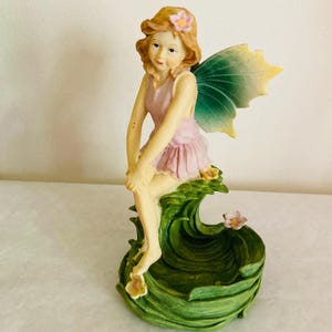 May include: A figurine of a seated fairy with blonde hair and a pink dress, adorned with a flower. The fairy has green and yellow wings and sits atop a green, leaf-like base. The figurine is a decorative item.