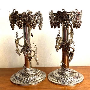 May include: A pair of silver candlestick holders with a grapevine design. The holders are tall and slender, with a round base and a decorative grapevine design that wraps around the stem. The grapevine design features leaves and grapes.