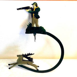 May include: A vintage metal sculpture depicting a hunter aiming a rifle at a deer. The hunter is dressed in a green jacket and hat, with tan pants. The deer is brown with large antlers. The sculpture is mounted on a curved, dark green metal base.