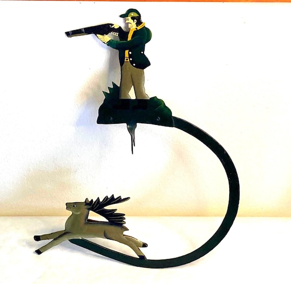 A hand-painted metal folk art balancing toy where a deer hunter defies gravity on a skyhook — charmingly weird, genuinely handmade Americana.