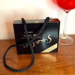 May include: A black wooden box purse with gold lettering that reads "Sinatra" and "My Way Limited Edition." The purse has a black braided strap and gold hardware. A glass of red liquid is in the background.