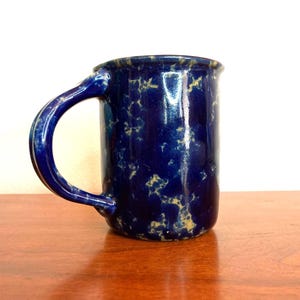 May include: A handmade ceramic mug with a dark blue glaze and a speckled yellow pattern. The mug has a large, curved handle and a cylindrical shape. The mug is sitting on a wooden surface.