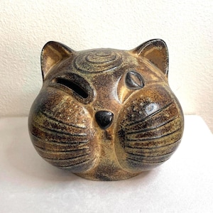 May include: A ceramic cat-shaped coin bank with a mottled brown and tan glaze. The feline figure features a rounded head, pointed ears, a small black nose, and incised lines for whiskers. A horizontal coin slot is positioned above the left eye.