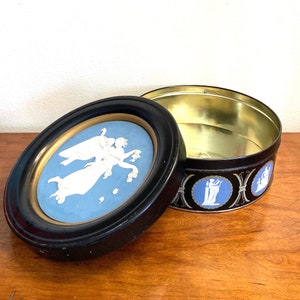 May include: A black and gold round tin with a blue and white cameo image of two women on the lid. The tin has a black frame around the cameo image. The tin is open and the inside is empty.
