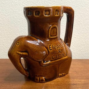 May include: Brown ceramic mug shaped like an elephant with a castle on its back. The castle has a small window and a brown glaze. The mug says "Elephant & Castle" on the front.