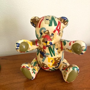 May include: A decorative teddy bear figurine with a glossy finish. The bear is cream-colored and covered in colorful letters, numbers, and small bear illustrations. The bear's paws and ears are green. The figurine is sitting on a wooden surface.