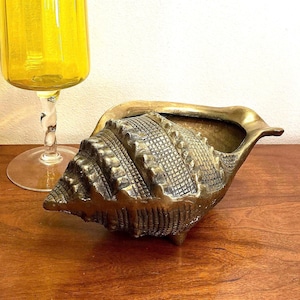 May include: A detailed, bronze-colored seashell-shaped decorative bowl. The bowl has a textured surface and is set on a wooden surface. A yellow glass with a twisted stem is in the background.