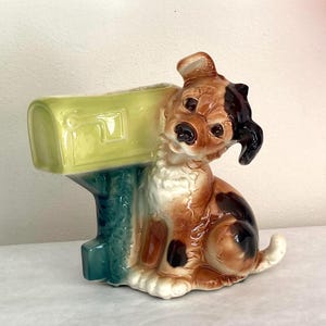 May include: A ceramic dog figurine with a mailbox. The dog is brown, black, and white, with a quizzical expression. The mailbox is light green and teal. The figurine is a decorative item.