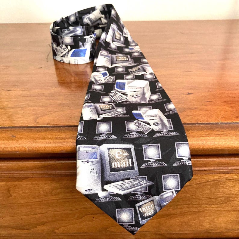 May include: A black necktie with a repeating pattern of computers, keyboards, and the words "e-mail" and "internet" in white. The tie is made of a smooth fabric and is folded over on a wooden surface.