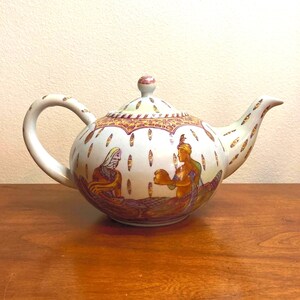 May include: White ceramic teapot with a hand-painted design of two figures in a scene with gold and red accents.