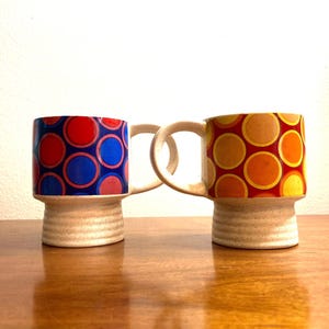 May include: Two ceramic mugs with a retro design. The mugs have a white base with a raised ring and a colorful pattern of red, blue, orange, and yellow circles.