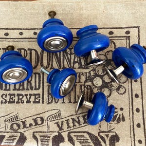 May include: Five blue ceramic cabinet knobs with silver metal accents. The knobs are round and have a raised center. They are arranged on a fabric surface with a pattern.