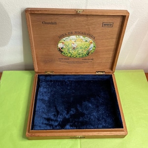 May include: Open wooden cigar box with a blue velvet interior. The lid features the text "Churchill" and a circular image with the words "Joya de Nicaragua." The box is labeled "Hand Made" and "Long Filler" with an "Imported" stamp.