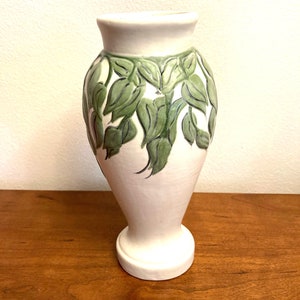 May include: White ceramic vase with a green leaf design. The vase has a rounded body and a flared base.