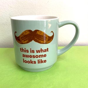 May include: A light green ceramic mug with a brown mustache graphic and the phrase "this is what awesome looks like" in red lettering. The mug has a curved handle and a white interior.